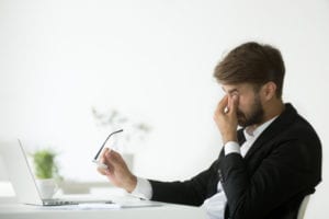Man at his desk holding his glasses and face due to a tension headache.