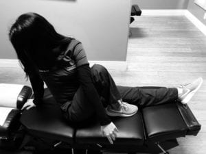 woman seated on chiropractic table with left leg outstretched and right leg pulled up with left arm crossed over right knee facing toward back wall