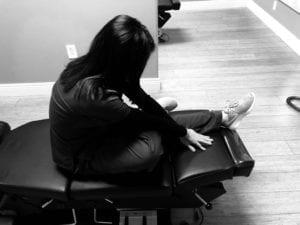 woman sitting on chiropractic table left leg outstretched with right leg crossed over left knee to stretch lower back and glutes