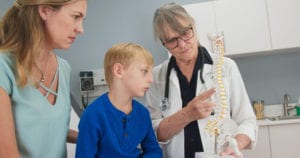 Senior woman pediatrician with spine model talking to little boy patient and his mother about scoliosis.